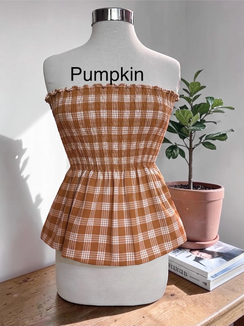 Peut inclure: Un haut bustier sans bretelles avec un motif &agrave; carreaux marron et blanc, dot&eacute; d'un corsage smock&eacute; &eacute;lastique et d'un bord sup&eacute;rieur volant&eacute;. Le mot Pumpkin est imprim&eacute; en noir au-dessus du v&ecirc;tement, pr&eacute;sent&eacute; sur un buste de mannequin blanc.