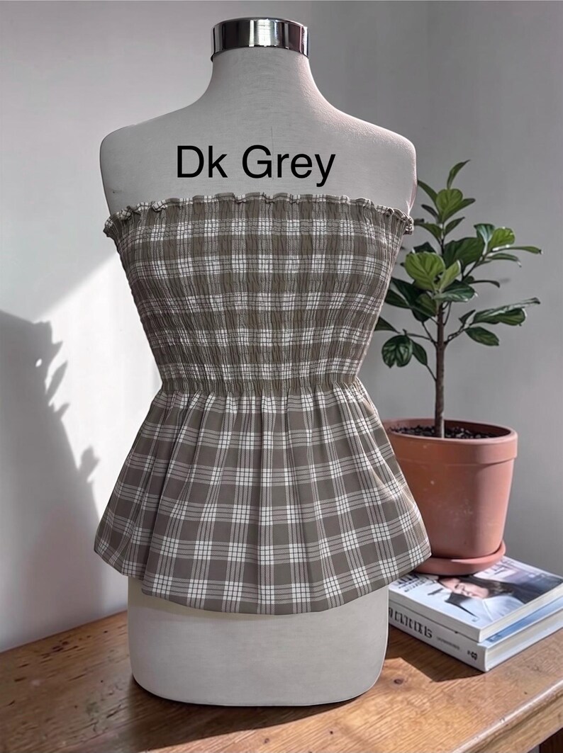 May include: A strapless tube top featuring a dark grey and white plaid pattern with a smocked, elasticized bodice and a flared peplum hem. The text Dk Grey is printed above the garment on the mannequin display.