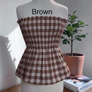 May include: A strapless peplum top featuring a brown and white plaid pattern with a smocked, elasticized bodice and a ruffled neckline. The word Brown is printed in black text above the garment, which is displayed on a mannequin torso.