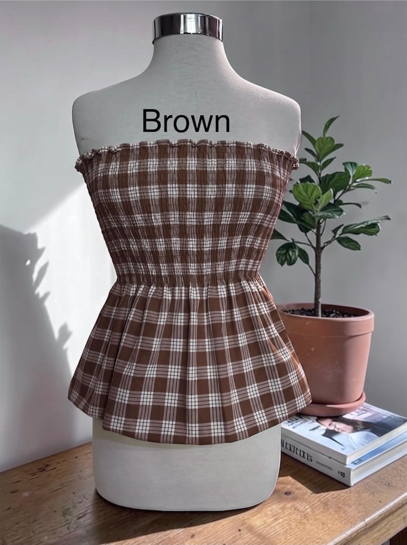 May include: A strapless peplum top featuring a brown and white plaid pattern with a smocked, elasticized bodice and a ruffled neckline. The word Brown is printed in black text above the garment, which is displayed on a mannequin torso.