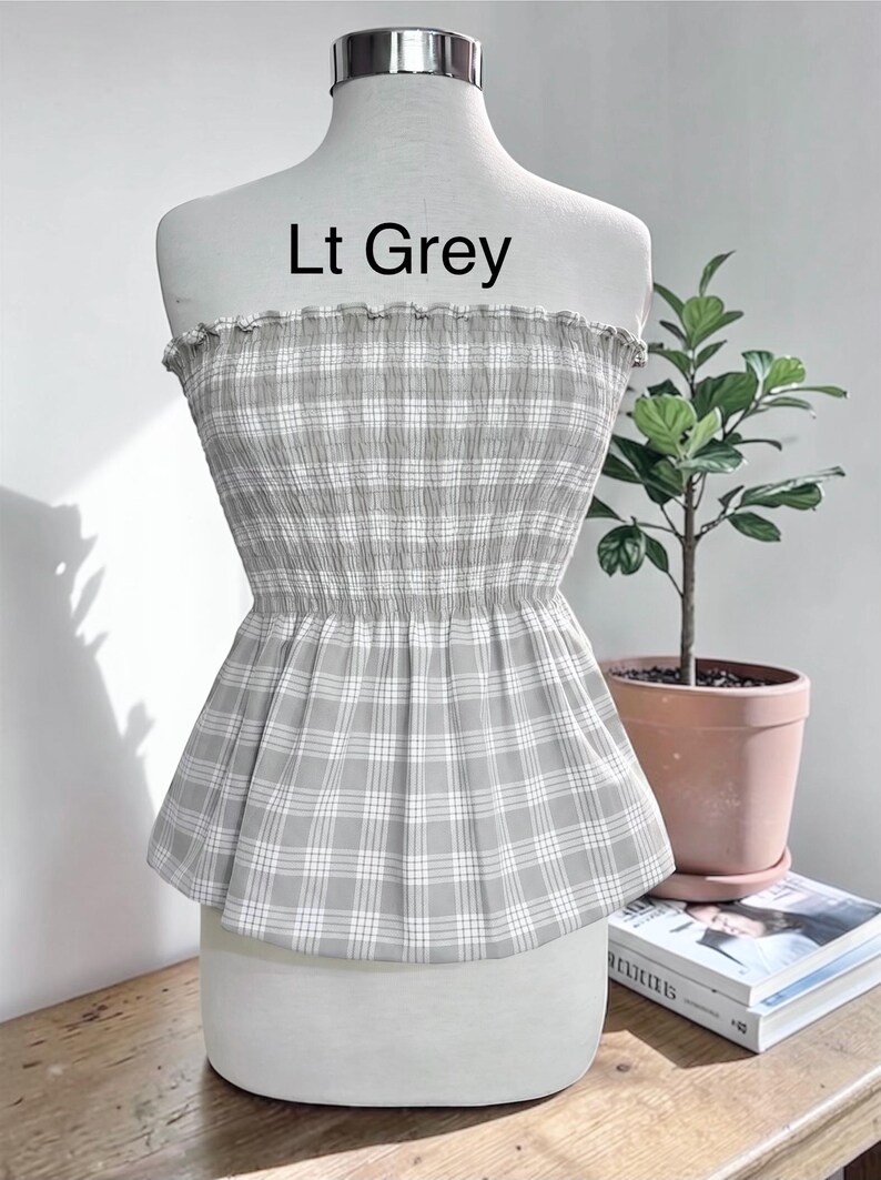 May include: A strapless tube top featuring a light grey and white plaid pattern with a smocked, elasticized bodice and a ruffled top edge. The garment is displayed on a mannequin and includes the printed text Lt Grey above the neckline.
