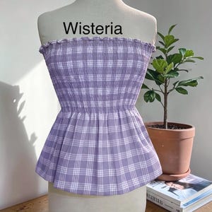 May include: A strapless tube top in a light purple and white plaid pattern. The garment features a smocked, elasticized bodice with a ruffled top edge and a flared peplum hem. The word Wisteria is printed in black text above the garment.