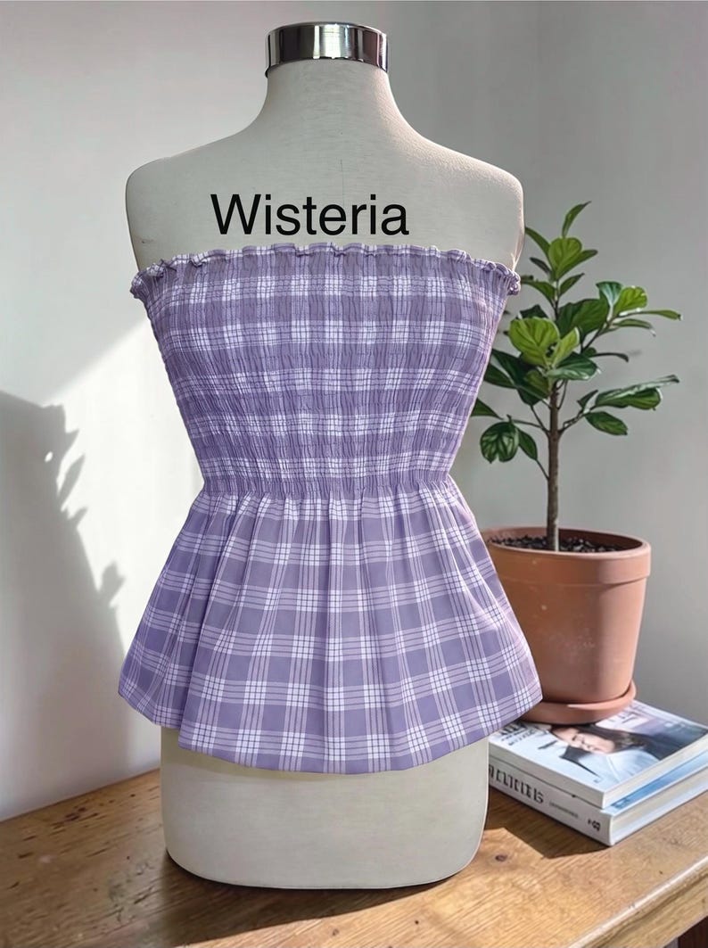 May include: A strapless tube top in a light purple and white plaid pattern. The garment features a smocked, elasticized bodice with a ruffled top edge and a flared peplum hem. The word Wisteria is printed in black text above the garment.