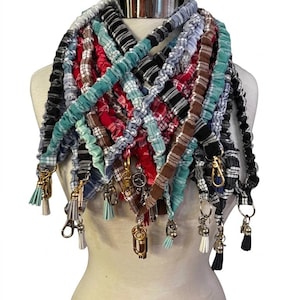 May include: A collection of multiple fabric lanyards featuring various plaid and striped patterns in colors including red, black, white, blue, and teal. Each lanyard has a gathered, elasticized texture and includes a gold-toned metal clasp and tassel.