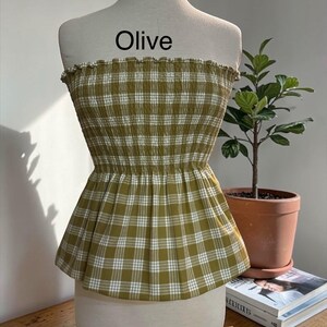 May include: An olive green and white plaid strapless top featuring a smocked, elasticized bodice with a ruffled top edge and a flared peplum hem. The word Olive is printed in black text above the garment on the white mannequin torso.