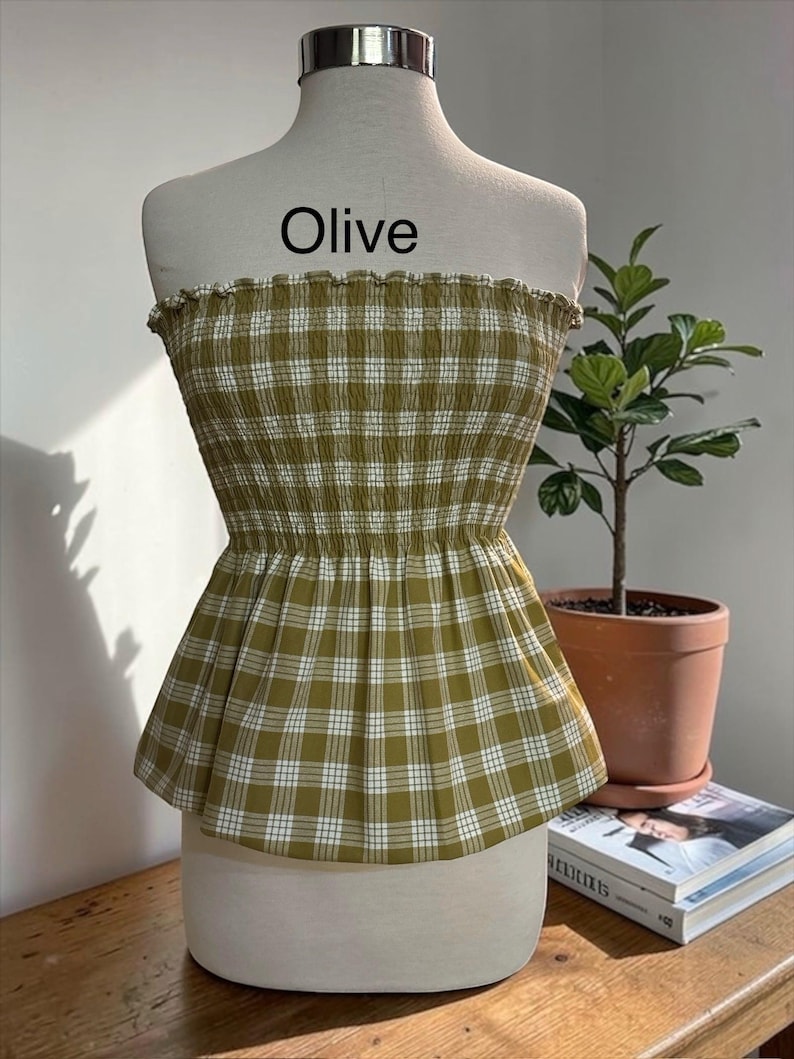 May include: An olive green and white plaid strapless top featuring a smocked, elasticized bodice with a ruffled top edge and a flared peplum hem. The word Olive is printed in black text above the garment on the white mannequin torso.