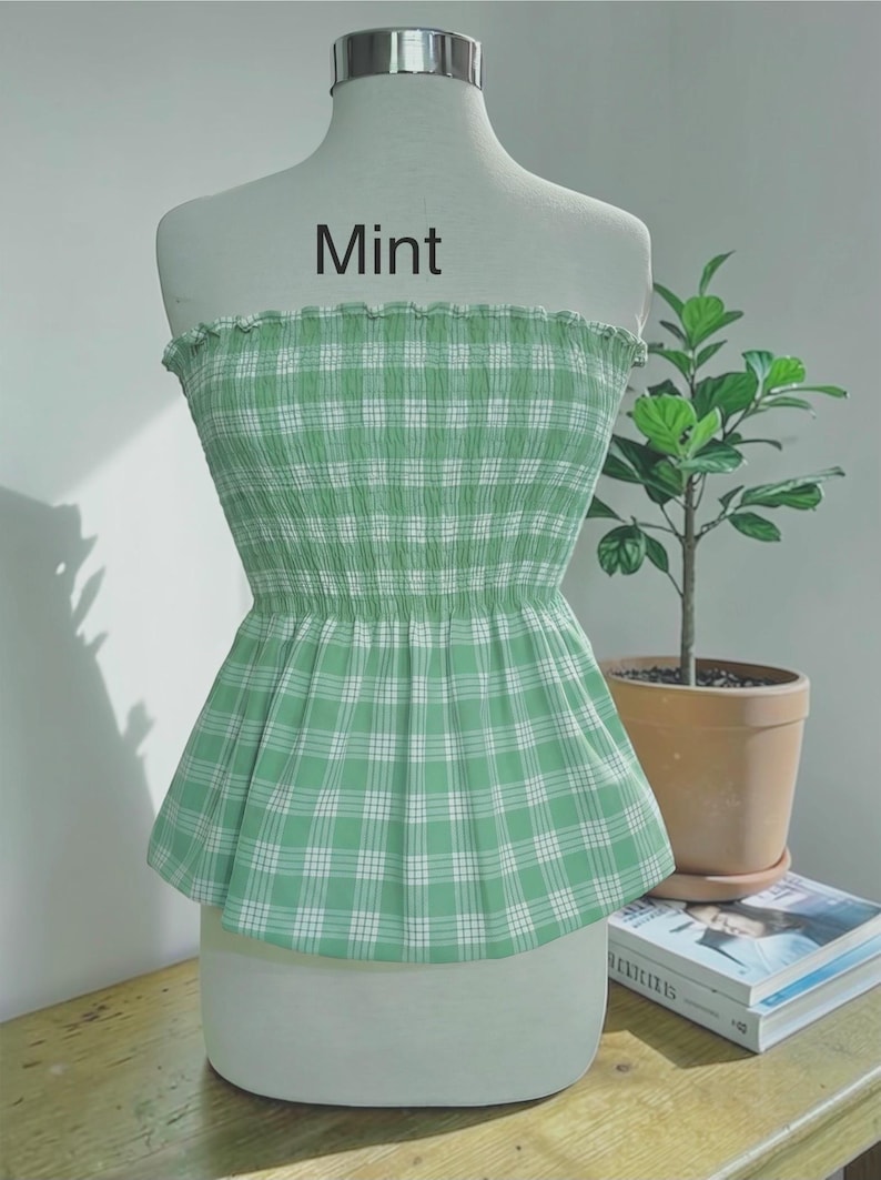 Peut inclure: Un haut bustier sans bretelles &agrave; motif &eacute;cossais vert menthe et blanc, dot&eacute; d'un corsage smock&eacute; &eacute;lastique et d'un bord sup&eacute;rieur volant&eacute;. Le mot Mint est imprim&eacute; en gris fonc&eacute; au-dessus du v&ecirc;tement sur le torse du mannequin blanc.
