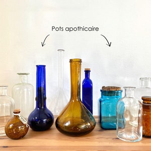 Può includere: Una collezione di vasi da farmacia di varie forme, dimensioni e colori, tra cui vetro trasparente, blu e ambrato. Alcuni vasi hanno tappi di sughero. Il testo "Pots apothicaire" è sopra i vasi.