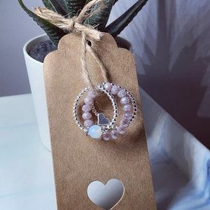 May include: Two beaded rings with a small silver heart charm and a white opal bead, presented on a brown tag with a heart-shaped cutout. The rings are light pink and silver, attached with twine to a small potted plant.