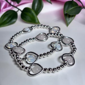 May include: Two silver beaded bracelets with heart-shaped charms. Each charm holds a round, light pink or white gemstone. The bracelets are displayed on a white surface with green leaves and pink fabric in the background.