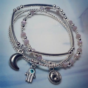 May include: Three silver bracelets with charms. The bracelets are layered and feature small, light purple beads. The charms include a crescent moon, a hamsa hand with a blue evil eye, and a yin yang symbol.