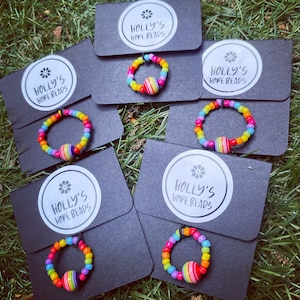 May include: Five black cardstock tags with white text that reads "HOLLY'S HOPE BEADS". Each tag has a rainbow-coloured beaded ring with a multi-coloured ball charm.