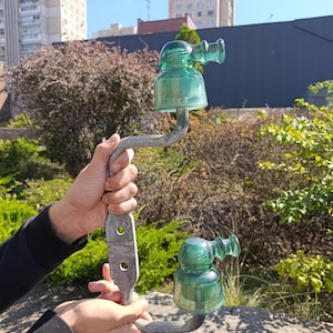 Puede incluir: Un soporte de metal vintage con dos aisladores eléctricos de vidrio verde unidos a su marco curvo. El brazo de metal gris y desgastado tiene dos orificios de montaje circulares y sostiene firmemente los componentes de vidrio verde.