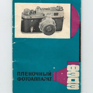 Op de afbeelding: Een vintage camerahandleiding met een blauwgroene omslag. De omslag toont een zwart-zilveren camera-afbeelding en tekst in het Russisch. Een magenta vorm bevindt zich aan de rechterkant.