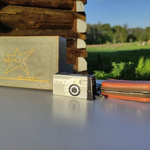 May include: A vintage silver camera, a brown leather case with a zip, and a grey box with a star logo and the text "Вега 2" are displayed on a table. The scene is set outdoors with a wooden structure in the background.