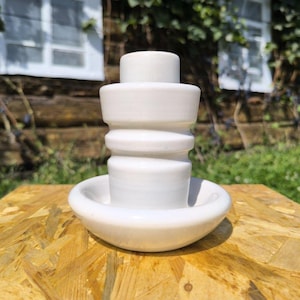 May include: A white ceramic candle holder with a distinctive, layered design. It has a cylindrical top, rounded sections, and a shallow, circular base. The piece is placed on a wooden surface, with a natural background visible.