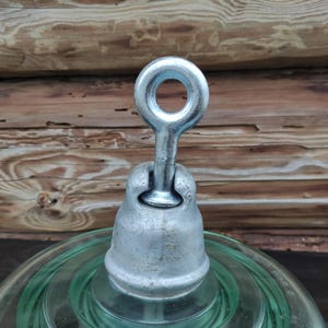 Op de afbeelding: Een vintage glazen isolator met een metalen dop en oogje. De isolator is helder, lichtgroen van kleur en de metalen componenten hebben een zilveren afwerking. De achtergrond is een houten oppervlak.