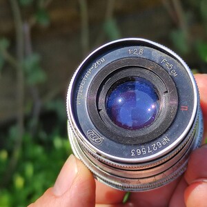May include: A vintage camera lens with a silver and black metal body. The lens has a blue glass element and markings including "1:2.8" and "F=5 cm". The serial number is visible. The lens is held against a blurred green background.