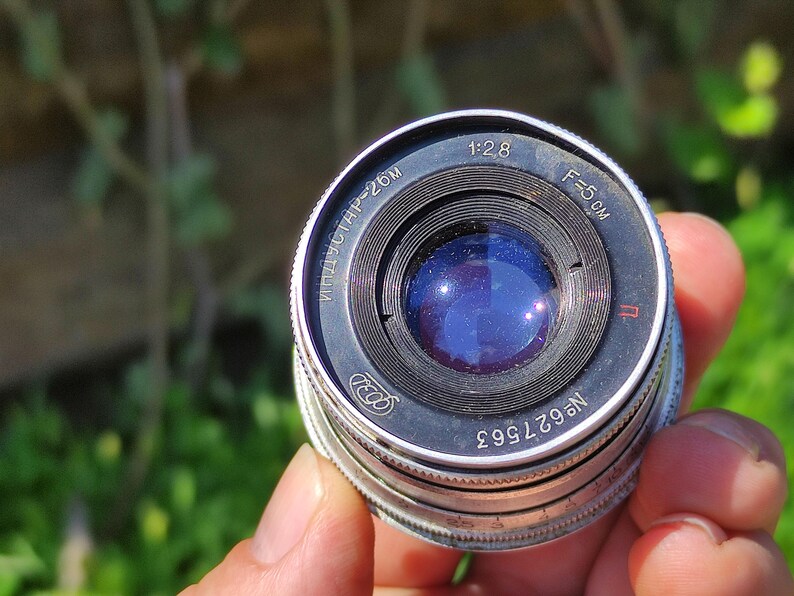 May include: A vintage camera lens with a silver and black metal body. The lens has a blue glass element and markings including "1:2.8" and "F=5 cm". The serial number is visible. The lens is held against a blurred green background.