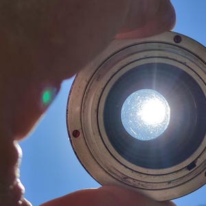 May include: A close-up of a silver lens with a dark interior, held against a bright blue sky. The sun shines through the lens, creating a bright, circular light.