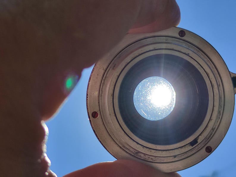 May include: A close-up of a silver lens with a dark interior, held against a bright blue sky. The sun shines through the lens, creating a bright, circular light.