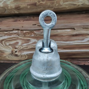May include: A vintage electrical insulator with a clear, green glass base and a silver-colored metal cap. The metal cap has a loop at the top and is attached to the glass base. The insulator is set against a rustic wooden background.