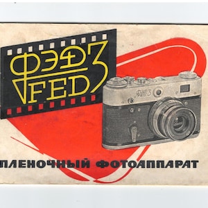 Puede incluir: Anuncio de cámara vintage con una cámara FED plateada y negra. El diseño incluye un gráfico de tira de película negra con texto amarillo y una forma de corazón roja. El texto en la parte inferior dice "ПЛЕНОЧНЫЙ ФОТОАППАРАТ".
