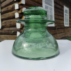 Puede incluir: Un aislador de vidrio verde vintage con una base redondeada y una parte superior acampanada. El aislador tiene un acabado liso y brillante y el número "1992" está grabado en el lateral. El objeto está sobre una superficie blanca.