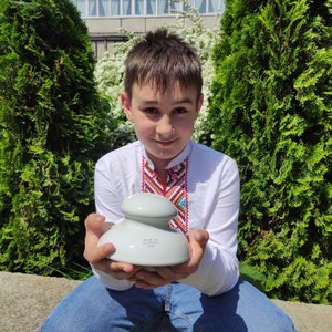 Op de afbeelding: Een jong persoon draagt een wit geborduurd shirt en houdt een keramische isolator vast met de tekst "Made in Ukraine 1983" erop.