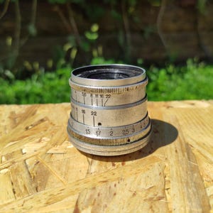 May include: A vintage silver camera lens with a black interior. The lens features multiple rings with engraved numbers and markings. It rests on a light brown wooden surface, with a blurred green backdrop.