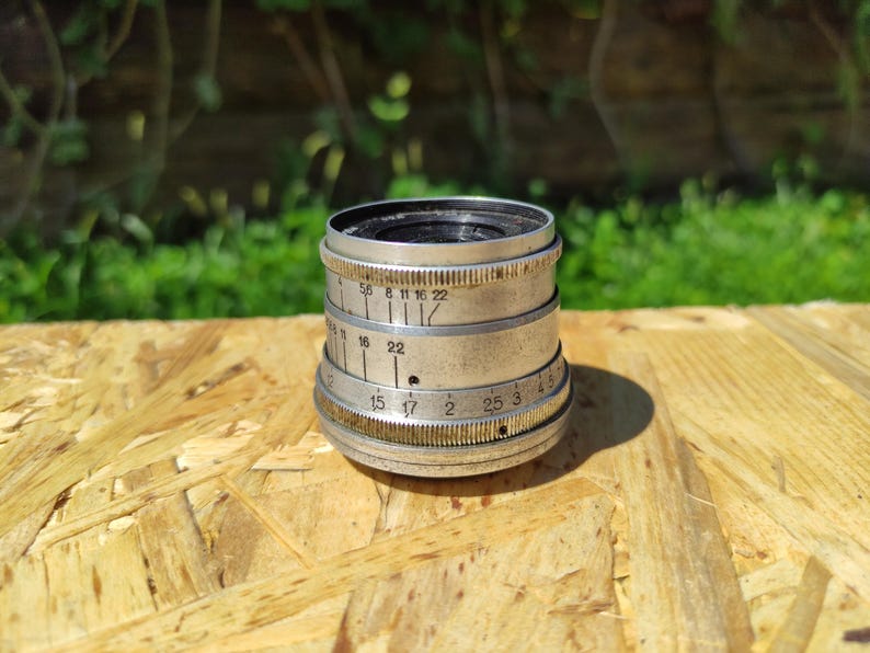 May include: A vintage silver camera lens with a black interior. The lens features multiple rings with engraved numbers and markings. It rests on a light brown wooden surface, with a blurred green backdrop.