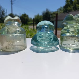 May include: Three vintage glass electrical insulators arranged in a row. The left and right insulators are clear with a slight green tint, featuring embossed markings including the number 1937. The centre insulator is a vibrant aqua blue.