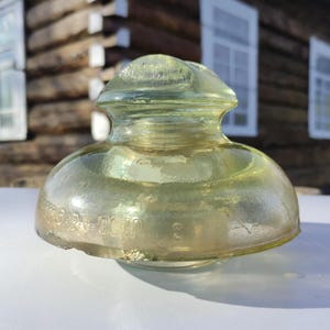 Può includere: Un isolatore in vetro verde chiaro vintage con una parte superiore e inferiore arrotondate. L'isolatore ha una superficie strutturata e sembra essere fatto di vetro spesso. La base ha testo in rilievo e il numero 3.