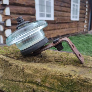May include: A vintage glass insulator with a metal bracket, resting on a moss-covered stone. The insulator is clear with a rounded top and a black rubber base. The bracket is rusty brown, and the background shows a wooden building.