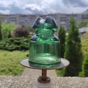 May include: A vintage green glass insulator with a rounded base and a double-grooved top. The insulator is mounted on a metal stand. The text "LUCENT VERRE" is visible on the glass. The background features greenery and a building.
