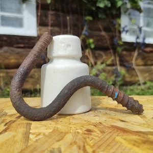 May include: A white ceramic insulator with a curved, rusty metal bolt. The bolt has a threaded end and a blue paint mark. The items are on a wooden surface, suggesting a vintage or industrial theme.