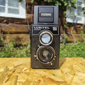 May include: A black Lubitel 166B Universal camera with a square body and two lenses. The camera has the word "LOMO" on top and "LUBITEL UNIVERSAL 166" on the front. It sits on a light brown wooden surface, with a blurred background of greenery and a wooden structure.