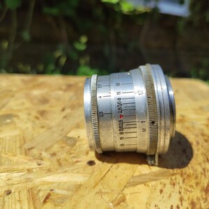 May include: A vintage silver camera lens with measurement markings. The lens has a ridged focus ring and a series of numbers and lines indicating aperture settings. The lens is set against a wooden surface.