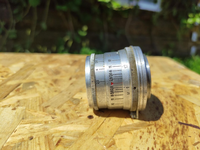 May include: A vintage silver camera lens with measurement markings. The lens has a ridged focus ring and a series of numbers and lines indicating aperture settings. The lens is set against a wooden surface.