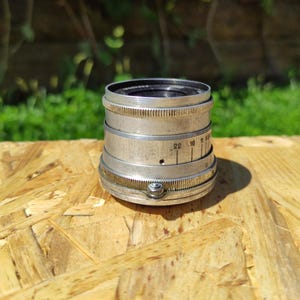 May include: A vintage silver camera lens with a black interior. The lens features a series of rings with numbers and markings. The lens is on a light brown wooden surface.
