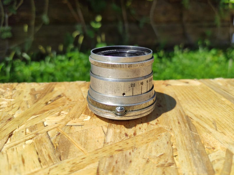 May include: A vintage silver camera lens with a black interior. The lens features a series of rings with numbers and markings. The lens is on a light brown wooden surface.