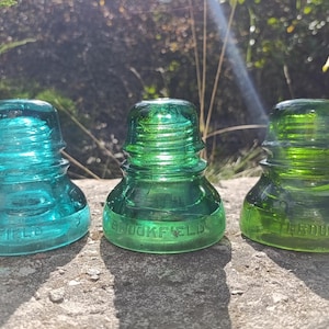 May include: Three glass electrical insulators arranged in a row. The left insulator is a light teal blue, the middle is a bright emerald green, and the right is a darker forest green. Each piece features the embossed text BROOKFIELD on the base.
