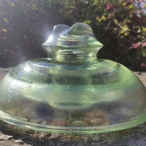Könnte beinhalten: Ein Vintage-Glasisolator in hellem Grün mit abgerundeter Basis und strukturierter Oberseite. Das Glas hat einen leichten Schimmer. Der Isolator liegt auf einer Steinoberfläche.