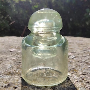 Könnte beinhalten: Ein klarer, alter Glasisolator mit abgerundeter Oberseite und zylindrischem Sockel. Das Glas hat einen leichten Grünstich und einige Unvollkommenheiten. Der Sockel des Isolators hat ein gestempeltes Design und die Nummer "1975".