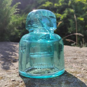 Op de afbeelding: Een turquoise glazen isolator met een afgeronde bovenkant en een cilindrisch lichaam. Het woord "HSMALL" is in reliëf op de zijkant aangebracht. De isolator staat op een stenen oppervlak, met een vage groene achtergrond.