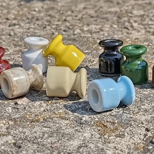 Może przedstawiać: Kolekcja dziesięciu małych, szpulowych izolatorów ceramicznych w różnych kolorach, w tym bordowym, białym, żółtym, czarnym, zielonym, jasnobrązowym, jasnoniebieskim i marmurkowym beżu. Każdy element posiada centralny otwór oraz rozszerzane krawędzie.