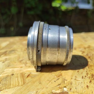 May include: A vintage silver camera lens with ridged edges and a series of rings. The lens is cylindrical and appears to be made of metal. The lens is resting on a wooden surface.