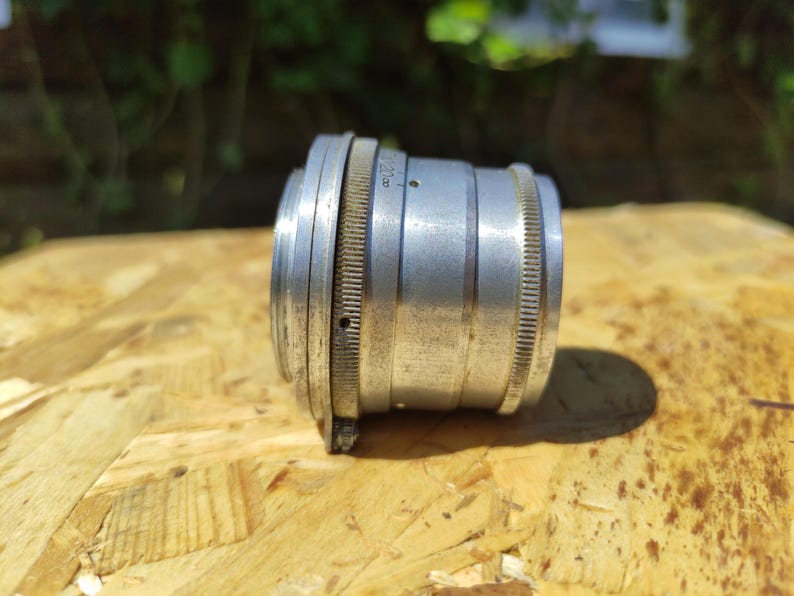 May include: A vintage silver camera lens with ridged edges and a series of rings. The lens is cylindrical and appears to be made of metal. The lens is resting on a wooden surface.