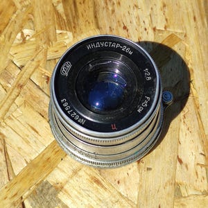 May include: A vintage camera lens with the text "ИНДУСТАР-26M" and "1:2.8 F=5CM" printed on the black and silver body. The lens has a blue reflective interior and is set against a wooden background.