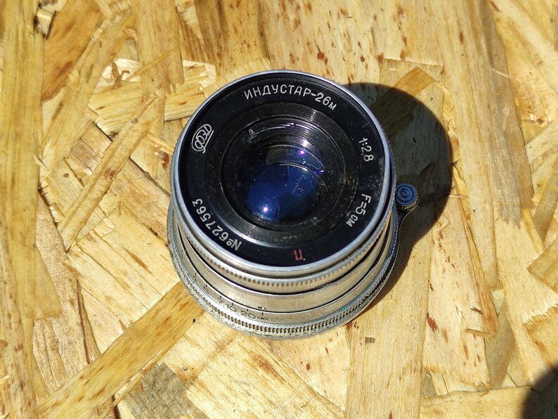 May include: A vintage camera lens with the text "ИНДУСТАР-26M" and "1:2.8 F=5CM" printed on the black and silver body. The lens has a blue reflective interior and is set against a wooden background.